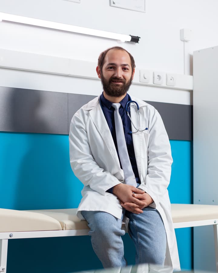 A smiling doctor in a white coat sitting on an exam bed in a bright clinic room