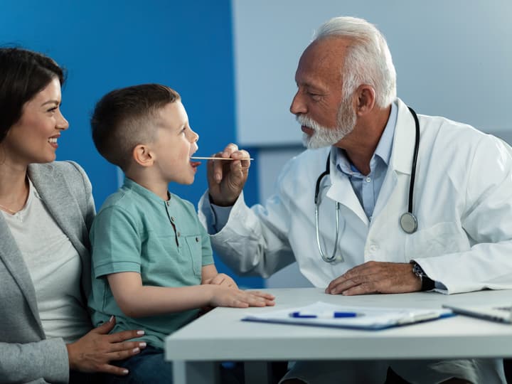 A doctor checking a young boy’s throat while his mother sits beside him